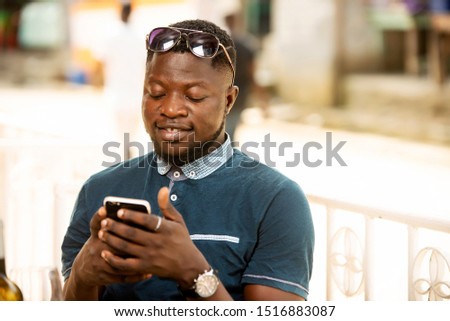 handsome african man sitting at an outdoors holding his phone in hand while happy while handling wearing glasses on his forehead looking at the screen of his phone