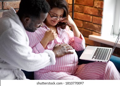 handsome african man stroke wife's pregnant tummy while she is sitting with laptop, love and expecting baby concept - Powered by Shutterstock