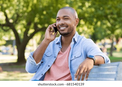 Handsome african man talking on phone at park. Young black man talking on cellphone while sitting on bench outdoor. African american smiling guy in a happy conversation at mobile phone.