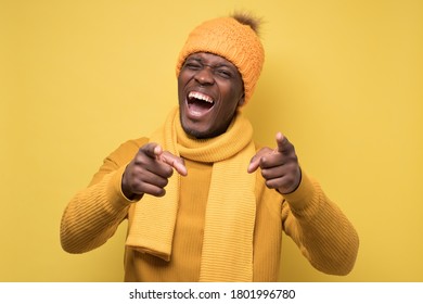 Handsome african man in yellow sweater and warm hat pointing at camera on joke. Studio shot