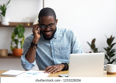 Handsome african manager sitting at office desk in front of laptop hold mobile phone make pleasant business or informal call. Successful businessman looking at financial statistic shown on documents