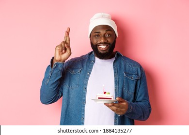 Handsome african-american guy celebrating birthday, making wish with fingers crossed, holding bday cake with candle, standing against pink background
