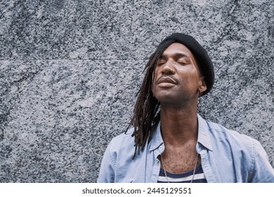 handsome African-American man in braids with stylish beret looking away while standing against grey wall and eyes closed - Powered by Shutterstock