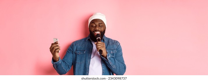Handsome african-american man singing karaoke, reading lyrics on smartphone app and holding microphone, standing over pink background.