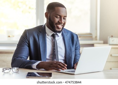 Handsome Afro American businessman in classic suit is using a laptop and smiling while working in office