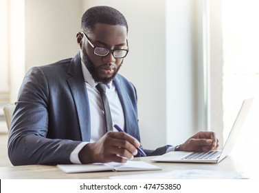 Handsome Afro American businessman in classic suit and eyeglasses is using a laptop and making notes while working in office - Powered by Shutterstock