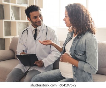 Handsome Afro American doctor in white coat is consulting beautiful pregnant woman in his office