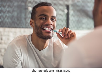 Handsome Afro American man is brushing his teeth while looking into the mirror in bathroom