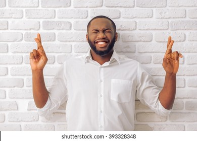 Handsome Afro American man in classic shirt is keeping fingers crossed, standing against white brick wall - Powered by Shutterstock