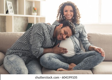 Handsome Afro American man and his beautiful pregnant wife are sitting on couch and smiling while spending time together at home