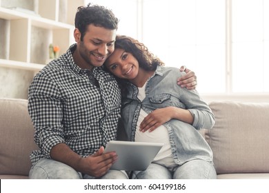 Handsome Afro American man and his beautiful pregnant wife are using a digital tablet and smiling while spending time together at home