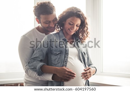 Handsome Afro American man and his beautiful pregnant wife are hugging and smiling while standing near the window at home