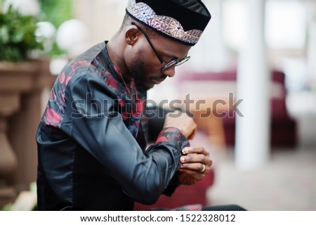 Handsome afro american man wearing traditional clothes, cap and eyeglasses in modern city sitting at couch.