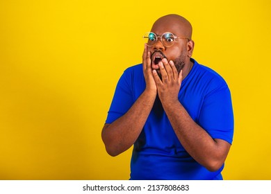 handsome afro brazilian man wearing glasses, blue shirt on yellow background. Amazed, startled, surprised, amazement, fear, wow, hands on cheek, mouth open,