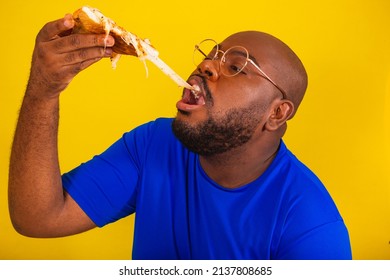 Handsome Afro Brazilian Man Wearing Glasses, Blue Shirt Over Yellow Background. Eating Pizza Slice, Cheese Stretching, Falling Cheese, Lots Of Cheese, Delicious, Creamy. To Stretch.