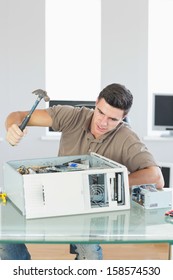 Handsome angry computer engineer destroying computer with hammer in bright office