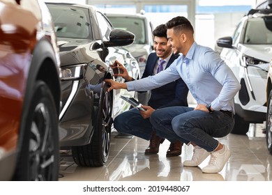 Handsome Arab Guy Customer Choosing New Sports Car In Showroom, Sitting By Luxury Black Auto And Having Conversation With Cheerful Sales Assistant, Touching Automobile Tire, Panorama