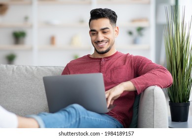 Handsome Arab Guy Surfing On Internet While Chilling At Home, Sitting On Couch And Using Modern Laptop, Copy Space. Cheerful Middle-eastern Man Reading Blog Or Looking For Job Online