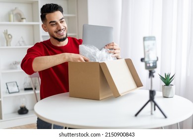 Handsome Arab Male Blogger Unboxing Parcel With Laptop On Camera, Smiling Middle Eastern Man Taking Computer Out Of Box While Recording Video Content For Tech Blog At Home, Using Smartphone On Tripod