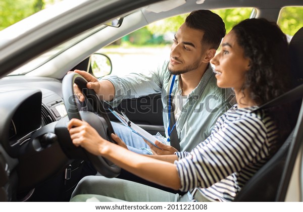 Handsome arab man driving instructor holding\
steering wheel, showing concentrated how to drive on the road,\
brunette lady young woman having lesson at driving school, side\
view, closeup