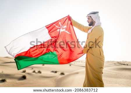 Handsome arabian man with traditional dress in the desert holding Oman flag