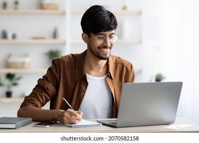 Handsome arabic guy employee surfing on Internet and taking notes, sitting at worktable in front of laptop, office interior, copy space. Unemployed young man looking for job online