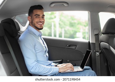 Handsome arabic guy in formal outfit entrepreneur sitting at auto back seat, using laptop with blank screen, typing on keyboard, sending emails while going to business meeting, mockup, copy space