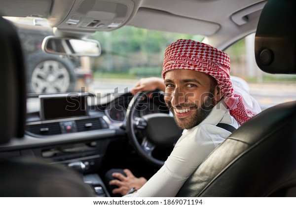 Handsome arabic man looking back smiling at\
passengers in the rear seat of his ehailing taxi cab. Arabian man\
wearing traditional headscarf\
keffiyeh