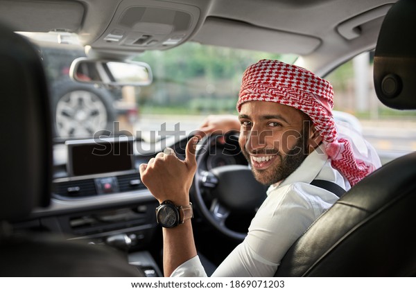 Handsome arabic man looking back\
smiling thumbs up at passengers in the rear seat of his ehailing\
taxi cab. Arabian man wearing traditional headscarf\
keffiyeh