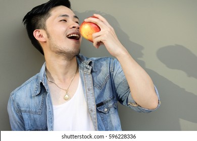 Handsome Asia Guy Love To Eat Fruit Especially Apple, It Is Good For Health. Charming Man Feel Happy And Relaxed. Attractive Man Hold Apple And Eating It. He Wear Denim Jacket And Vest. Copy Space