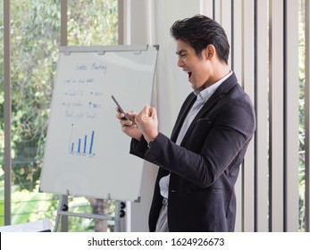 Handsome Asian businessman standing by the window uses the internet and social media on his smartphone to work in the office.He is happy because job is successful.Garden background with green trees. - Shutterstock ID 1624926673