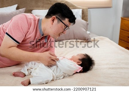 Handsome Asian father wearing pink shirt, Lying and teasing with his baby daughter, on a soft brown bed, The father gently invites the little boy to talk, and cherishes his daughter's hand.