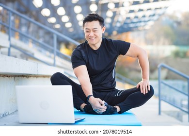 handsome asian fitness coach holds workout classes online using video call webcam and laptop. sitting outside in a modern stadium. Pilates training or stretching. Physical exercises. Records Lessons - Powered by Shutterstock