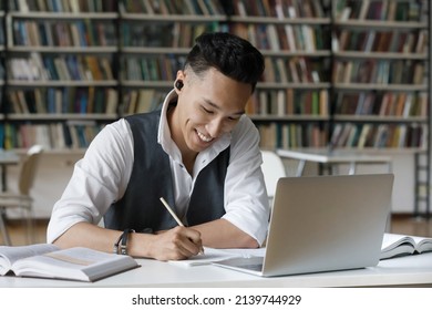 Handsome Asian guy hold pencil take notes in copybook while studying alone in library. Student wear earbuds listen audio task, makes assignment sit at table. Education, improve knowledge, tech concept
