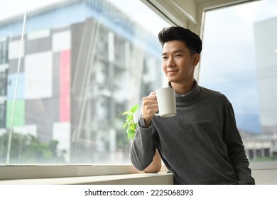 Handsome Asian Male Drinking Coffee Standing Near Office Window Overlooking Cityscape