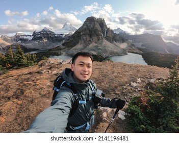 Handsome Asian Male Traveler Taking Selfie Portrait On The Hill With Mount Assiniboine In National Park At BC, Canada