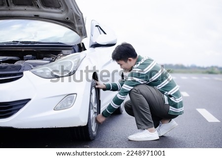 Handsome Asian man checking the punctured tyre on his car loosening the nuts with a wheel spanner before jacking up the vehicle in street. Men hold rubber to repair. Change tire. broken car concept.