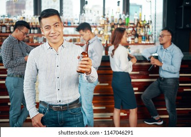 Handsome Asian Man Holding Beer Bottle In Outstretched Hand Smiling At Camera With Friends In Bar On Background