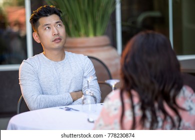 Handsome Asian Man On An Outdoor Date With A Black Female.  The Couple Are Sitting In A Restaurant Or Cafe Setup For Speed Dating.  He Looks Bored And Disappointed.