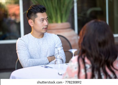 Handsome Asian Man On An Outdoor Date With A Black Female.  The Couple Are Sitting In A Restaurant Or Cafe Setup For Speed Dating.  He Looks Bored And Disappointed.