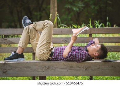 Handsome Asian man is reading a book lying on bench with one leg crossing on his knee