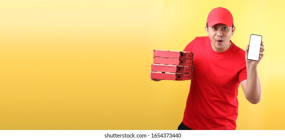  Handsome Asian Man In Red Cap, Giving Food Order Italian Pizza In Cardboard  Boxes Isolated On Yellow Background In Studio With Copy Space. Holding Mobile Phone With Blank  White  Empty Screen. 