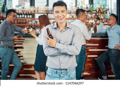 Handsome Asian Man In Shirt Holding Glass Bottle Of Beer And Smiling At Camera In Bar With Colleagues