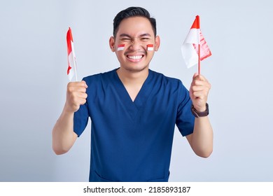 Handsome Asian Man Wearing Blue Male Nurse Uniform Holding Indonesian Flag And Celebrating Indonesian Independence Day August 17 Isolated On White Background Studio Portrait