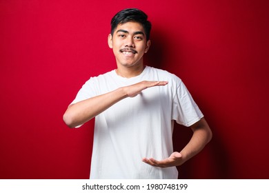 Handsome Asian Man Wearing White Tshirt Over Red Background Gesturing With Hands Showing Big And Large Size Sign, Measure Symbol. Smiling Looking At The Camera. Measuring Concept.