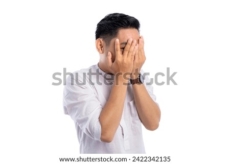 Handsome Asian Muslim man peeking at the camera through his fingers, embarrassment covering his face isolated on white background. Ramadan and Eid Fitr celebration concept