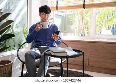 Handsome Asian Teenager Man Wearing Casual Clothes In Glasses Using Smart Phone Browsing Internet On Mobile Phone Device At The Living Room 