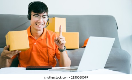 Handsome Asian Thai Male Employee Is Contacting Customer Via Laptop Webcam To Sell In The Packaging Box.Online Order Confirmation Working At Home In Orange Uniforms.Ecommerce Internet Work From Home.