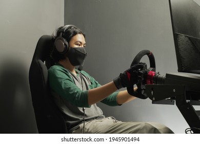 Handsome Asian Young Man Gamer Wearing Mask In Headphones With Pc Computer Playing Car Racing Video Game At Home And Steering Wheel