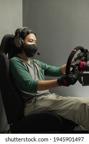 Handsome Asian Young Man Gamer Wearing Mask In Headphones With Pc Computer Playing Car Racing Video Game At Home And Steering Wheel